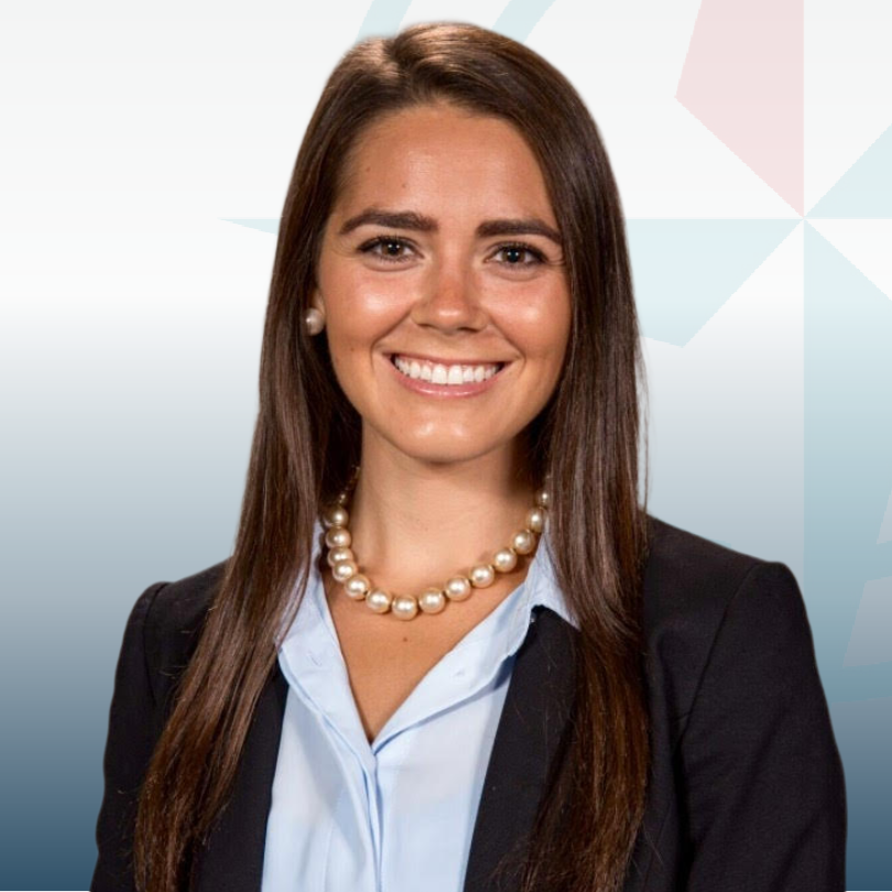 Smiling woman with long brown hair, wearing a black blazer, light blue shirt, and pearl necklace. Background features geometric shapes.