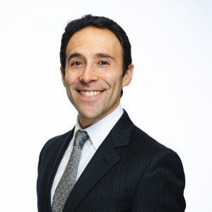 Smiling man wearing a pinstripe suit and patterned tie against a plain white background. The mood is professional and approachable.
