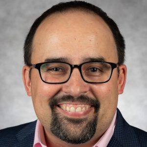 Smiling man with glasses, dark hair, and a goatee, wearing a checkered suit and pink shirt against a light gray background, conveying a friendly tone.