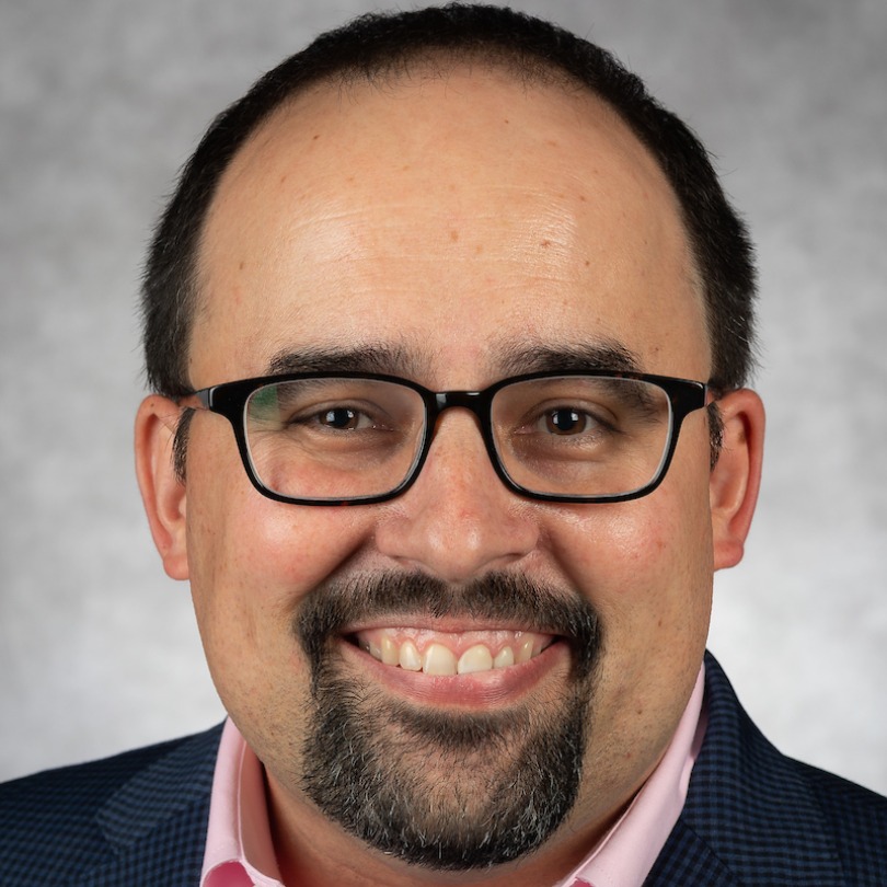 Smiling man with glasses, dark hair, and a goatee, wearing a checkered suit and pink shirt against a light gray background, conveying a friendly tone.