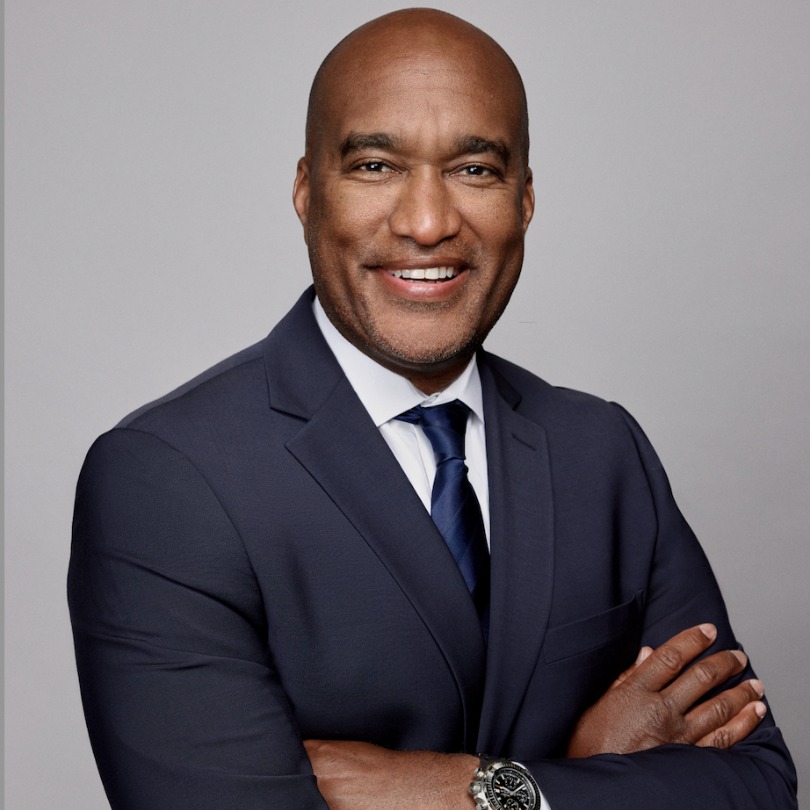 Smiling man in a navy suit with a white shirt and dark tie, arms crossed. Gray background suggests professionalism and confidence.