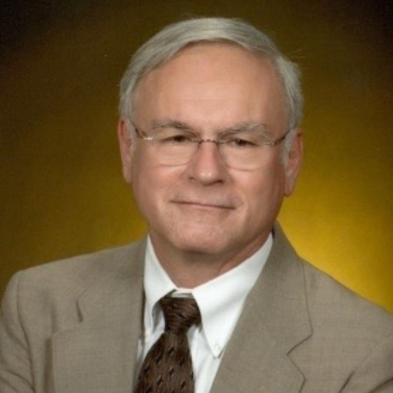Elderly man in a beige suit and brown tie with glasses, smiling softly against a warm, golden-brown background. Professional and approachable tone.
