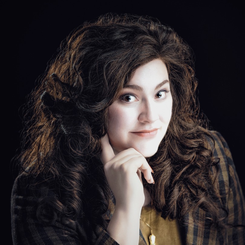 Young woman with long, curly hair smiles softly, resting her chin on her hand. She's wearing a plaid jacket against a dark background, conveying warmth.