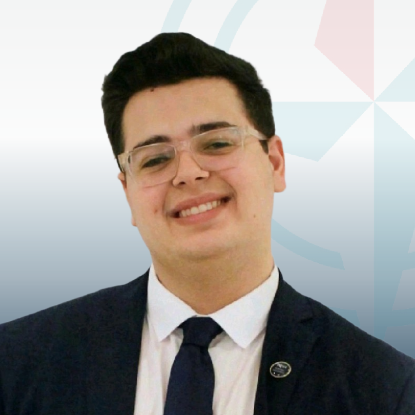 Smiling man in glasses and suit, dark hair, against a light blue geometric background. He conveys a cheerful and professional tone.